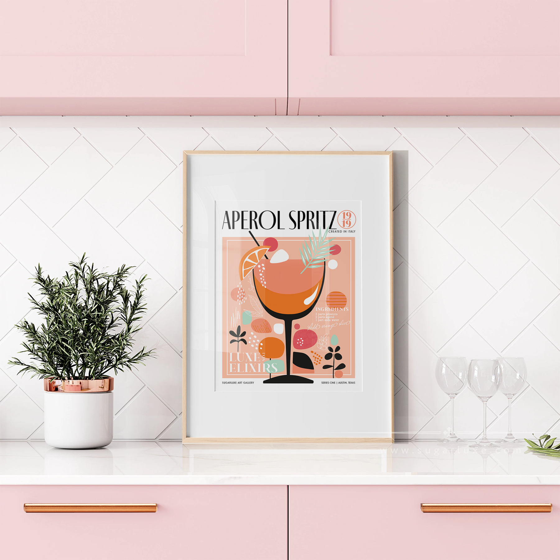 pink kitchen art aperol spritz mid century modern design cocktail posters Framed vintage retro poster with a cocktail illustration on a kitchen countertop with a potted plant and glasses. The poster features the words "Aperol Spritz" at the top in bold, black letters. It depicts a stylized illustration of a cocktail glass filled with a vibrant orange liquid, garnished with a cherry and a slice of citrus. The background is pink with decorative circular and foliage motifs in shades of mint green, peach, and coral. The poster is set against a kitchen wall with white tiles arranged in a herringbone pattern. Above the tiles, there are pink cabinets with rose gold handles. To the left of the poster, there is a small potted plant with dark green leaves in a white pot with a rose gold rim. To the right, there are three empty wine glasses placed on the countertop.